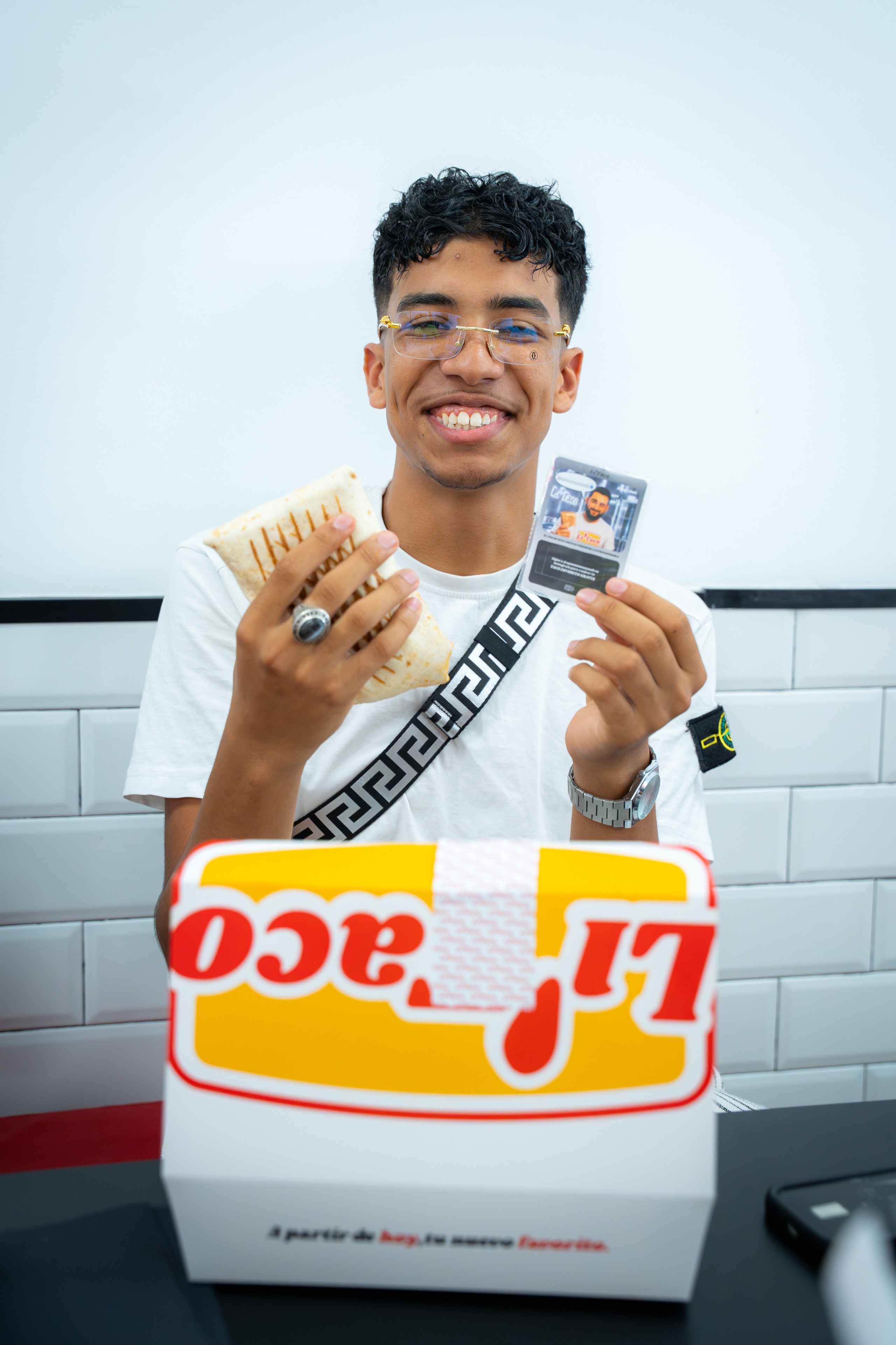 Un chico sonríe con un taco y un cromo en la mano, tras una caja LiTaco