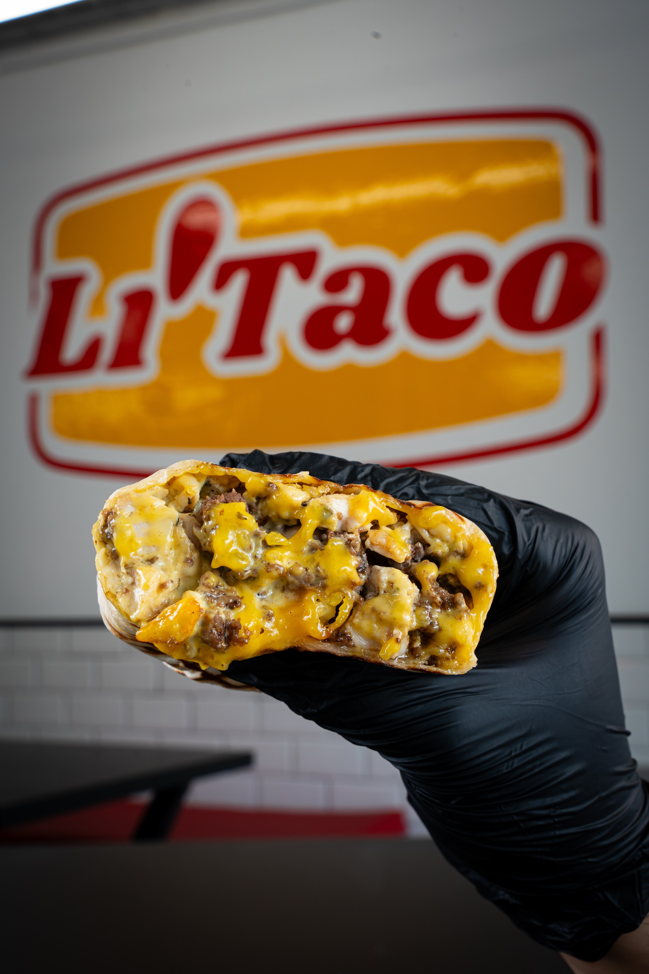 Mano con guante negro sostiene un taco abierto, con el rótulo de LiTaco detrás