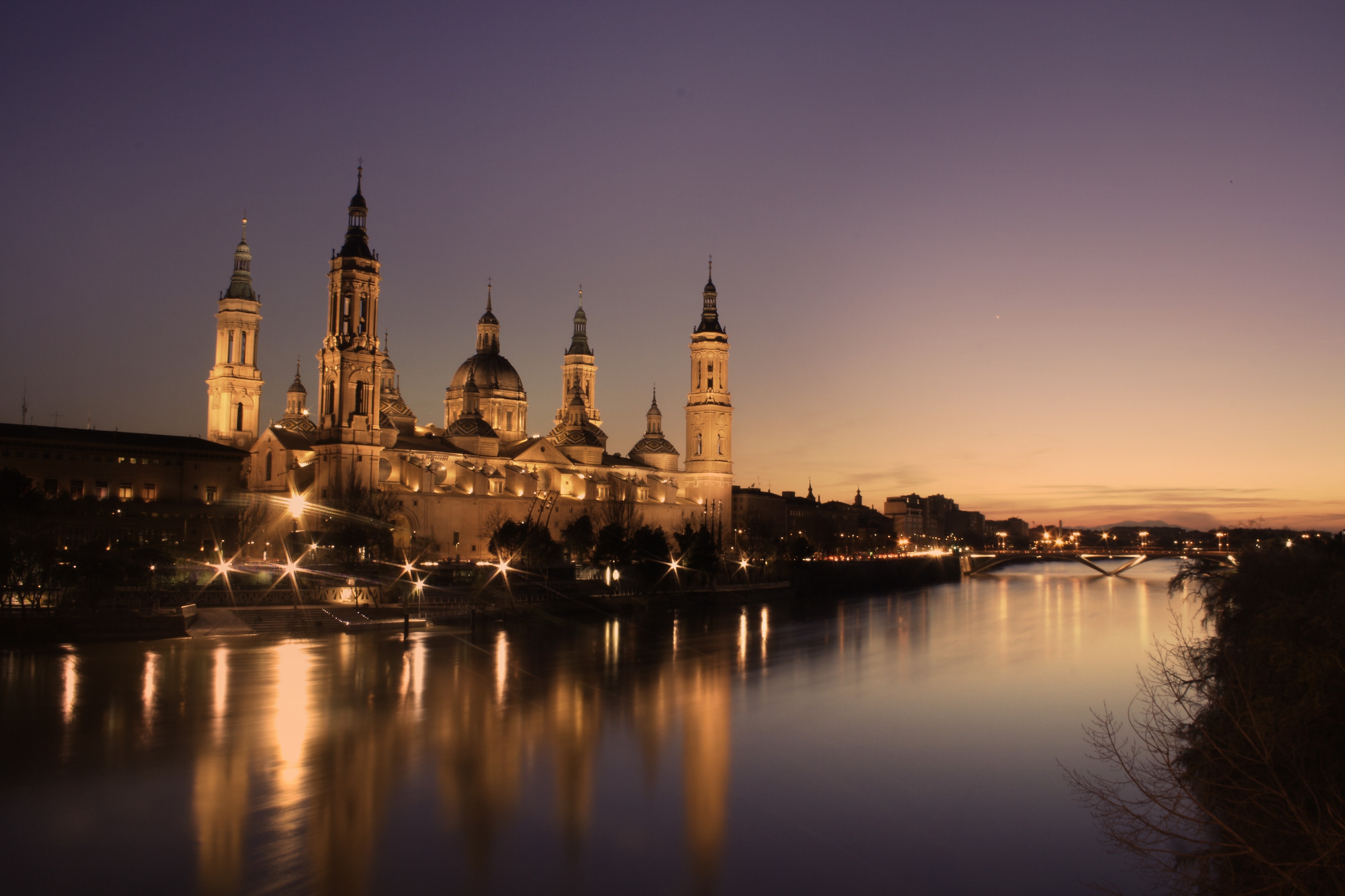 La basílica del Pilar iluminada al atardecer, reflejada en el Ebro bajo un cielo naranja y morado